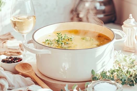 A large white pot filled with vegetable broth is simmering on the stove, releasing steam. Fresh herbs float on top next to a glass of white wine and bowls of spices, creating a warm atmosphere.