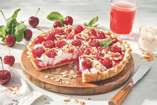 A delicious cherry tart sits on a wooden board, topped with whipped cream, almonds, and fresh cherries. A glass of pink drink accompanies this dessert, creating a vibrant summer treat.