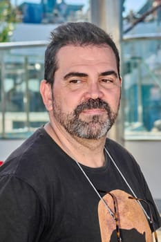 A man with a black t-shirt and glasses hangs around his neck, looking sideways in a casual pose in an urban outdoor environment.