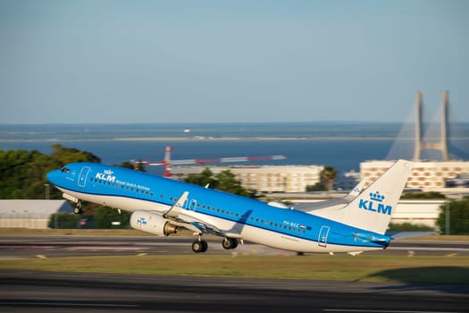 Lisbon, Portugal - Apr 3, 2024: Panning shot of KLM Royal Dutch Airlines aircraft Boeing 737-800 take off runway at Lisbon airport on sunny day, clear sky, Vasco da Gama bridge in background. Vacations, getaway, travel destination, blurred background