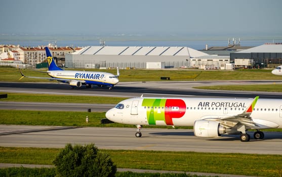 Lisbon, Portugal - Apr 3, 2024: TAP or Air Portugal airplane and Ryanair aircraft taxiing on runway at Lisbon Airport on sunny day. Airport hangars and city buildings in background. Busy travel destination, low-cost airline, national airport