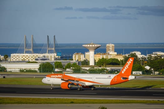 Lisbon, Portugal - Apr 3, 2024: EasyJet low-cost airline Airbus A321 neo aircraft taxiing on runway at Lisbon airport on sunny day, clear sky, Vasco da Gama bridge in background. Vacations, weekend getaway, summer travel destination, takeoff, landing