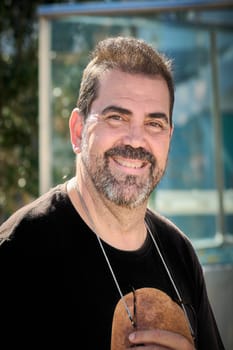 A man wearing a black t-shirt smiles warmly at the camera, standing outdoors with a soft urban background and natural lighting.
