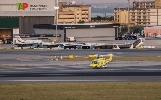 Lisbon, Portugal - May 9, 2024: INEM, national institute of medical emergency, yellow rescue helicopter ready to take off on taxiway at Lisbon airport, TAP Maintenance and Engineering hangar, buses, urban buildings in background at golden hour