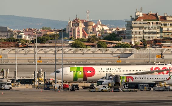 Lisbon, Portugal - Apr 24, 2024: TAP Air Portugal aircrafts waiting for boarding by airport hangars on sunny day. City landscapes in background, clear blue sky, summer haze, travel destination, vacations, transportation, Portuguese travel hub.