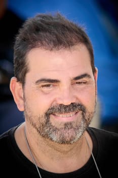 A casually dressed man wearing black lowers his gaze slightly while smiling, framed by a softly blurred outdoor urban scene.