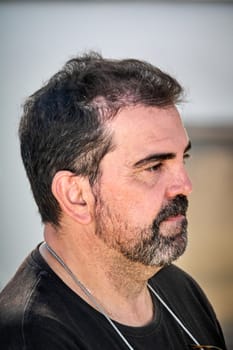 A man in a black shirt and a delicate chain necklace gazes thoughtfully to his side, standing outdoors in a serene urban setting.