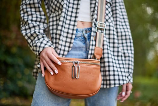 A young woman with wavy brown hair and blue eyes confidently poses outdoors in a lush autumn park. She wears a trendy black-and-white checkered shirt over a white crop top and classic blue jeans, accessorized with a tan leather crossbody bag with a striped strap. The soft natural lighting and blurred background of green foliage create a dreamy and cozy fall atmosphere. This fashionable portrait is ideal for lifestyle branding, autumn fashion campaigns, social media promotions, and advertising seasonal collections.
