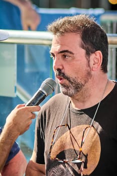 A casually dressed man in black t-shirt listens attentively during an outdoor interview, with a microphone close to his face in a blurred urban setting.
