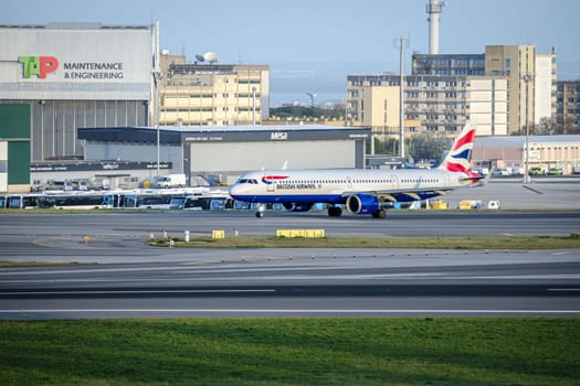 Lisbon, Portugal - Apr 3, 2024: British Airways airplane Airbus A321neo taxiing on runway at Lisbon Airport. TAP Maintenance and Engineering hangar, infrastructure, buses, city buildings in background. Clear blue sky, sunny day, travel destination