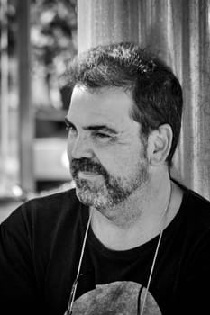 A man with a faint smile wears a black T-shirt, captured from a slightly elevated angle, standing outdoors with a blurred urban setting.