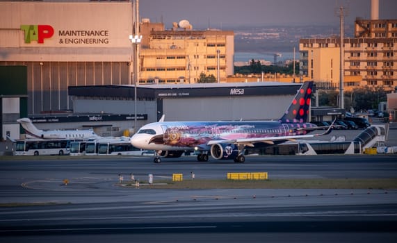 Lisbon, Portugal - May 9, 2024: Brussels Airlines Airbus A320 neo in colorful Amare livery inspired by Tomorrowland festival taxiing at Lisbon airport at golden hour. TAP Maintenance and Engineering hangar, buses, and urban buildings in background