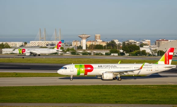 Lisbon, Portugal - Apr 3, 2024: Two TAP Air Portugal airline Airbus A321neo aircrafts ready for takeoff at Lisbon airport, clear blue sky, sunny day. Summer haze, vacation, travel destination, cityscape and Vasco da Gama bridge in background
