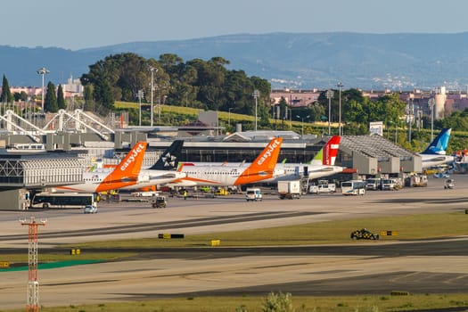 Lisbon, Portugal - Apr 24, 2024: TAP, Azores Airlines and EasyJet airplanes in Lisbon airport ready for boarding and takeoff on sunny day, clear blue sky. Ground support vehicles, airport Terminal 1 infrastructure, city buildings in background.