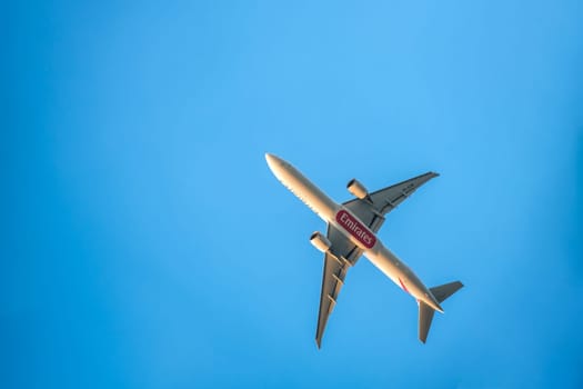 Lisbon, Portugal - Apr 3, 2024: Upshot of Emirates Boeing 777-300ER airplane flying overhead against clear blue sky on sunny day. Aircraft, airline, branding or logo, bottom fuselage, sunlight. Passenger long-haul flight, flag carrier from Dubai, UAE