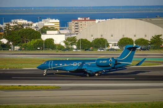Lisbon, Portugal - Apr 29, 2024: Sleek private jet taxiing on an airport runway, surrounded by urban buildings, greenery and a distant river view. Aircraft model Gulfstream G650ER, long-range business jet, private aviation, exclusive service