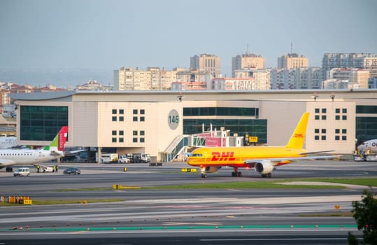 Lisbon, Portugal - Apr 3, 2024: DHL cargo airplane Boeing 757-200F taxiing at Lisbon Airport and TAP Air Portugal passenger airplane parked at nearby gate. Airport terminal, city skyline in background. International travel and shipment hub, sunny day