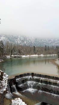 Tiered Waterfall Lechfall in a Snowy Mountain Landscape in Fuessen, Germany. A beautiful multi-tiered waterfall cascading over dark stone steps into a turquoise river Lech, surrounded by a snowy forest. The misty Alpine mountain in the background adds a mystical atmosphere to the winter scenery.
