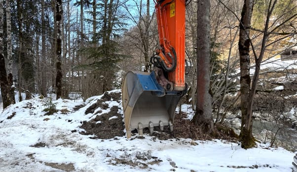 Orange Construction Excavator in a Winter Forest Clearing with Snow Cover. An orange construction excavator is parked in a snowy woodland, surrounded by trees and a beautiful winter landscape, showcasing industrial machinery blending into natural settings.