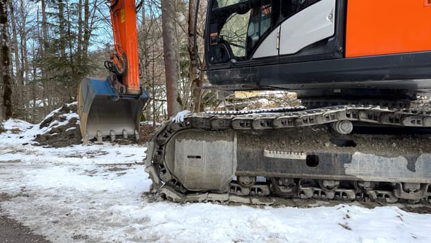Orange Excavator on Snowy Ground With Tracks and Mechanical Bucket in Forest. An orange excavator with caterpillar tracks and a mechanical bucket is on snowy terrain surrounded by trees. Perfect for themes of construction, heavy machinery, winter work, or industrial operations.