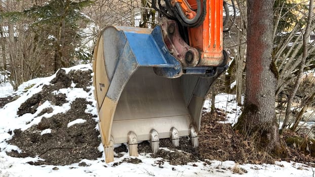 Construction Excavator Bucket by Forested Snow Landscape. Excavator bucket near forest during winter, showcasing industrial machinery and contrasting snowy environment. Useful for images depicting construction technology, industrial equipment, winter projects, or outdoor construction activities.