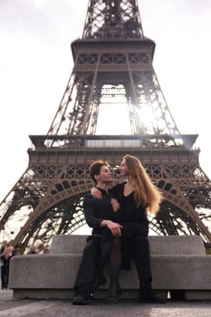 A romantic couple in black outfits sits on a bench near the Eiffel Tower, gazing into each other eyes as the golden sunlight shines through.