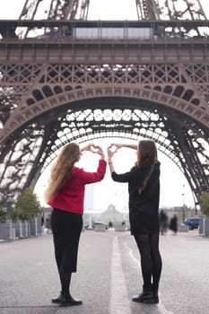 Two young women stand beneath the Eiffel Tower, forming a heart shape with their hands, symbolizing love, friendship, and adventure.
