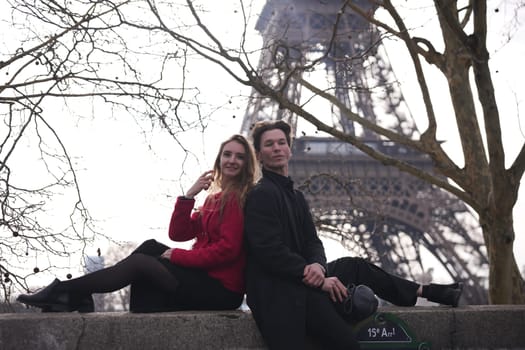A fashionable young couple leans back against each other on a stone ledge near the Eiffel Tower, embracing a relaxed Parisian vibe.