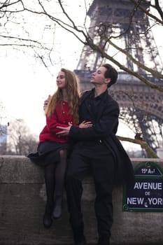 A young couple shares a romantic moment on Avenue de New York in Paris, sitting on a stone ledge with the Eiffel Tower behind them.