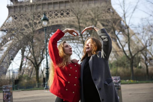 Two happy women stand in front of the Eiffel Tower in Paris, forming a heart shape with their hands, celebrating friendship and joy.