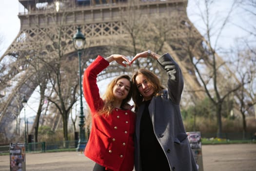 Two stylish friends pose in front of the Eiffel Tower in Paris, forming a heart shape with their hands, symbolizing love and friendship.