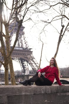 A young woman in a red coat relaxes on a stone ledge in Paris, with the Eiffel Tower creating a perfect backdrop.