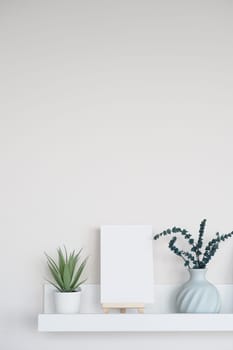 A clean shelf holds a blank canvas, green plants, and a vase.