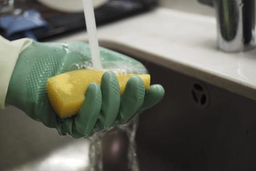 A person rinses a yellow sponge while wearing green gloves.