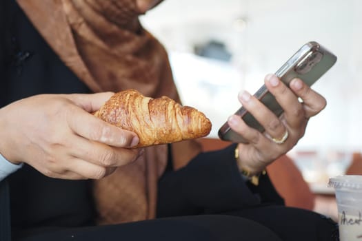 A person holds a croissant and looks at their phone in a cafe.