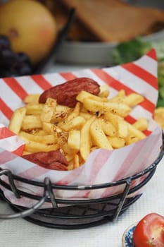 Fries served in a basket with sausage, surrounded by fresh produce and color.
