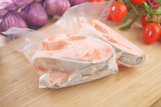 Frozen salmon fillets sit on a wooden board with fresh tomatoes and onions.