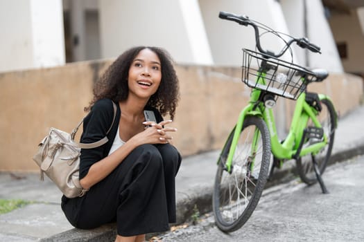 This image shows a woman joyfully using her phone while perched on a curb next to her bright green bicycle, representing a blend of technology and eco-conscious living.