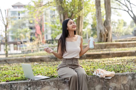 A joyful young woman enjoys a refreshing drink, embodying a sustainable lifestyle while sitting in a peaceful park with her laptop.