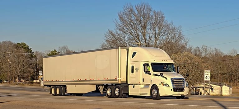 Freight liner Semi-Truck Driving on Open Highway in a Rural Area, CDL Truck America, semi truck and trailer travels along an open road under a blue sky in a rustic rural setting, showcasing commercial transportation, logistics efficiency, and professional trucking