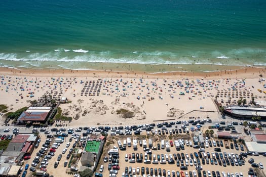 Aerial view of crowded beach with adjacent car parking area, showcasing turquoise sea and sunbathers enjoying the sunny day on the sandy shore