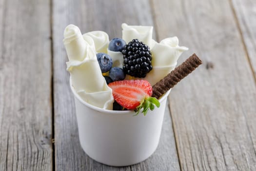 Delicious rolled ice cream in a cup, garnished with fresh berries, a chocolate wafer stick, and fruit toppings, served on a rustic wooden table