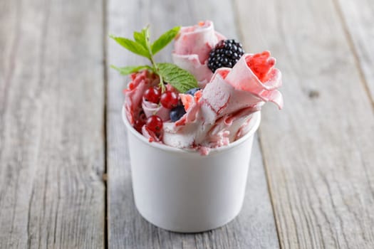 Delicious rolled ice cream in a cup, garnished with fresh berries, a chocolate wafer stick, and fruit toppings, served on a rustic wooden table