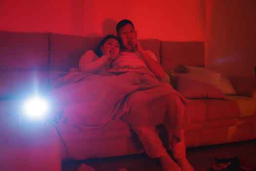 Asian couple relaxing on sofa under blanket, enjoying film using video projector and eating snacks in cozy red light living room