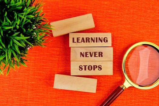 Wooden blocks display the phrase learning never stops surrounded by a magnifying glass and a small green plant on an orange surface