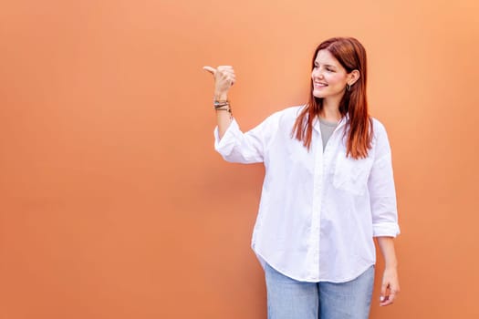 smiling young woman in white shirt and blue jeans is pointing to the side with the thumb, copyspace for text