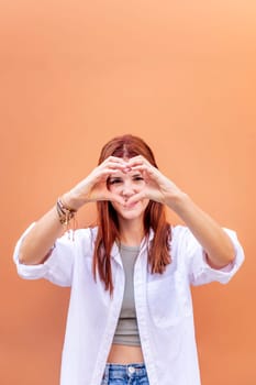young woman standing in front of orange wall doing a heart shape with hands while looking at camera, she is wearing a white shirt, concept of love, copyspace for text