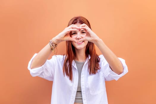 young woman standing in front of orange wall doing a heart shape with hands while looking at camera, she is wearing a white shirt, concept of love, copyspace for text