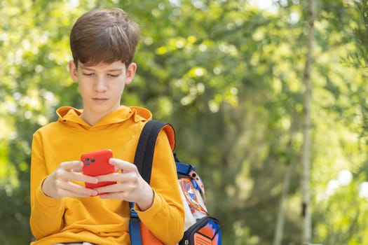A teenage student in a yellow hoodie sits in a park and looks at his smartphone. Green trees in the background create a relaxed outdoor setting. Concept of education, online learning, and technology.