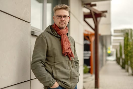 Young Attractive Man On A Street Wears Glasses And A Scarf, Looking Into The Camera